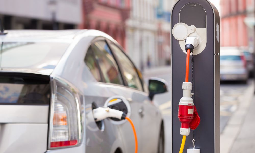 Gümüş renkli hibrit elektrikli bir araba, şehir caddesindeki bir şarj istasyonuna takılı. İstasyonda birden fazla priz bulunuyor ve araba turuncu bir kabloyla bağlanıyor. Binalar ve bir yol, modern kentsel sürdürülebilirliği sergileyen bir fon oluşturuyor.