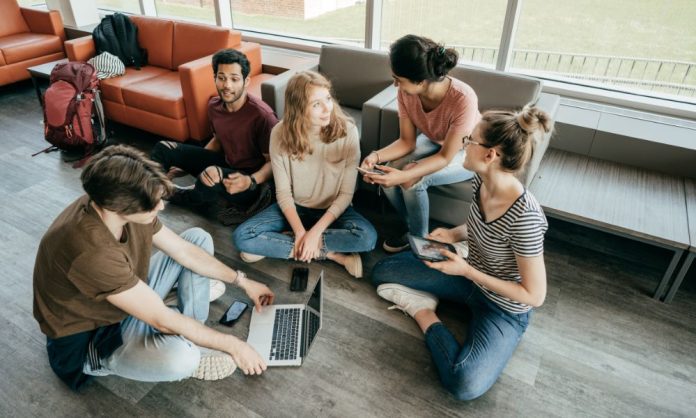 Beş genç yetişkinden oluşan bir grup, sosyal gelişim hakkında sohbet ederek yerde rahatça oturuyor. Bazıları tablet ve telefon tutarken, biri dizüstü bilgisayar kullanıyor. Kanepelerle çevrililer ve arka planda yumuşak bir ışık yayan büyük pencereler var.