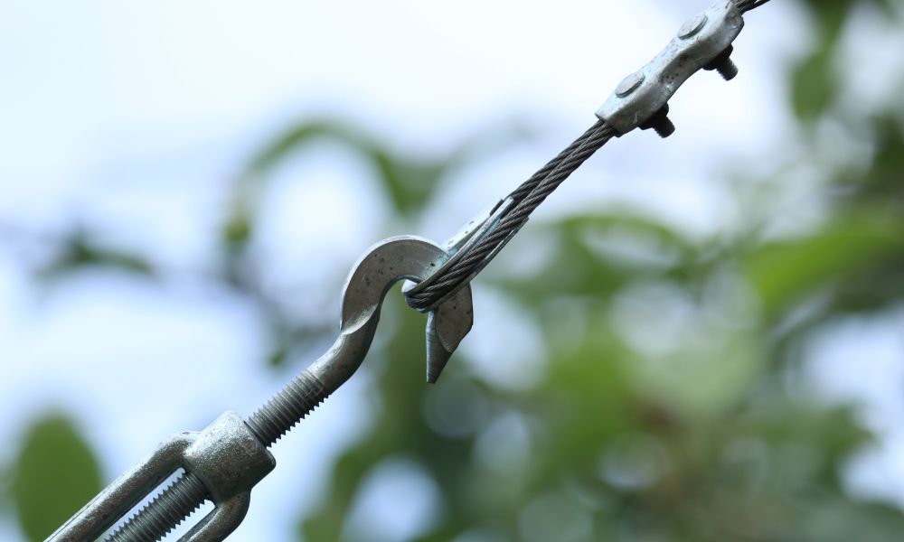 Çelik bir kabloya bağlı metal bir kancanın, bulanık doğal bir arka planla yakın çekimi. Kanca, çeşitli bağlantılarda direnci ve kabloyu sabitleyen kelepçeleri gösteren bir gerdirme sisteminin parçasıdır.