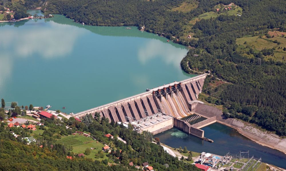 Bir tarafında turkuaz su ve diğer tarafında savak bulunan hidroelektrik enerji kullanan büyük bir barajın havadan görünümü. Yemyeşil tepeler ve dağınık binalarla çevrili, berrak gökyüzü suyun yüzeyine parlak bir şekilde yansıyor.