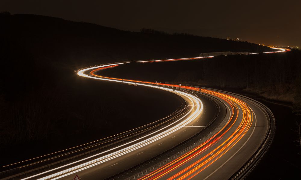 Gece vakti bir otoyolun uzun pozlama fotoğrafı, araçların ışık izlerini gösteriyor. Yol, karanlık, engebeli bir manzarada kıvrılıyor, arabaların kırmızı ve beyaz ışık çizgileri dinamik, parlayan bir yol oluşturuyor; sanki mükemmel bir uyum için görünmeyen bir hız sabitleyici tarafından yönlendiriliyormuş gibi.