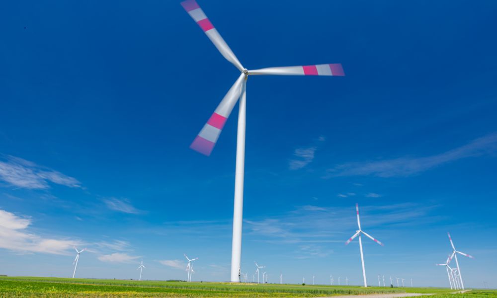 Büyük bir rüzgar türbini, kanatları açık mavi bir gökyüzünün altında yeşil bir alanda zarifçe dönerken kinetik enerjiyi kullanır. Enerjinin özünü yakalayan birkaç başka türbin arka planda görülebilir.