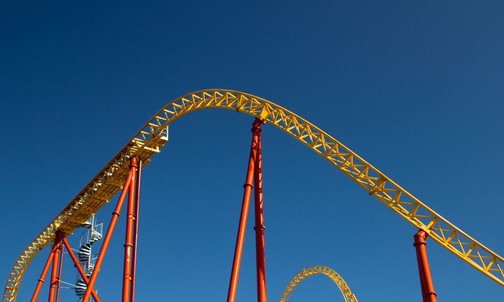 Açık mavi gökyüzünün önündeki parlak sarı ve turuncu renkli roller coaster pisti, kinetik enerjinin heyecan verici dinamiklerini sergilerken, görünür bir halka ve birkaç yüksek destek, arka planla çarpıcı bir kontrast oluşturuyor.