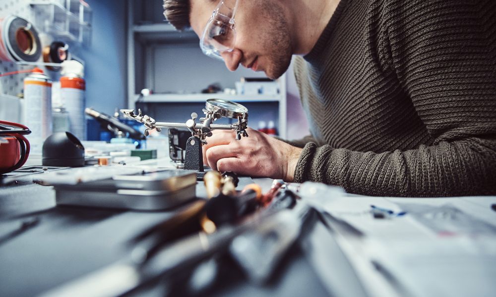 Güvenlik gözlüğü takan bir kişi, nano teknoloji alanında büyüteç ve hassas aletler kullanarak dağınık bir çalışma tezgahında elektronik bileşenleri birleştirmeye odaklanıyor. İyi aydınlatılmış atölyede çeşitli aletler ve malzemeler etrafa dağılmış durumda.