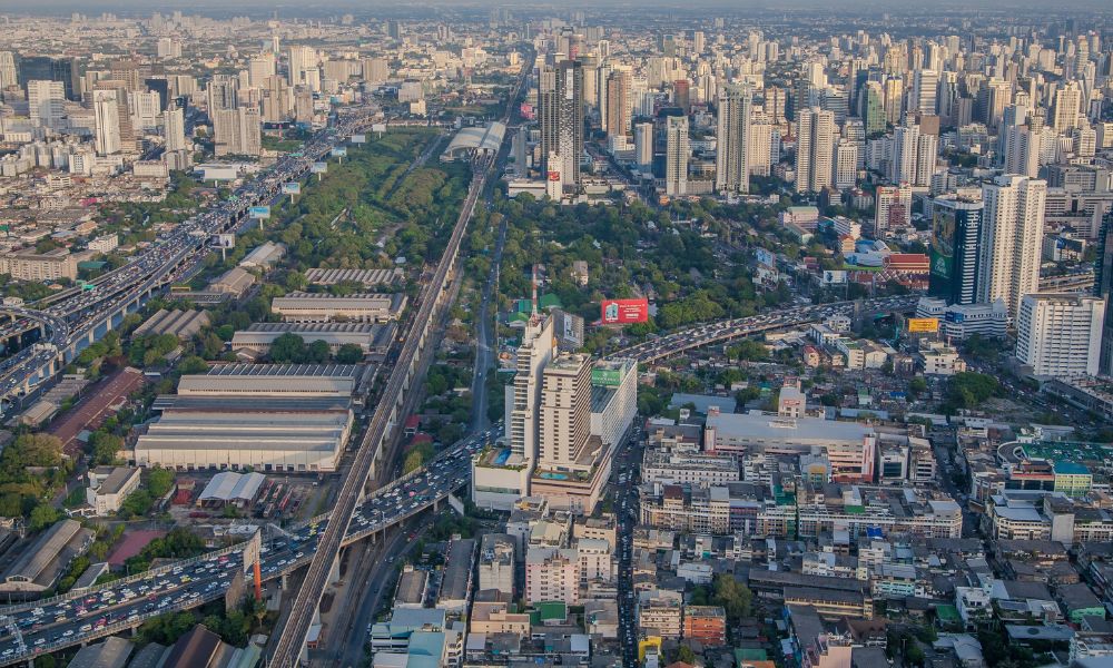 Hareketli bir şehir manzarasının havadan görünümü, yüksek katlı binalar, yoğun yollar ve yeşil alanların bir karışımını içeren kentleşmeyi sergiliyor. Açık bir gökyüzünün altında ufuk çizgisi uzağa doğru uzanırken ana yollarda trafik sıkışıklığı görülebiliyor.