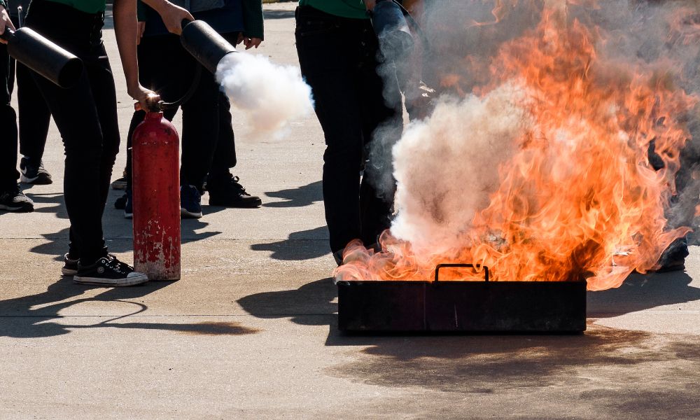 Bir yangın güvenliği tatbikatı sırasında, insanlar kontrollü bir açık alev üzerinde yangın söndürücüler kullanarak aktif olarak katılırlar. Ateş, metal bir kapta parlak bir şekilde yanarken, katılımcılar ona beyaz söndürücü bulutları yönelterek bu tür durumlarla başa çıkmada temel becerileri sergilerler.